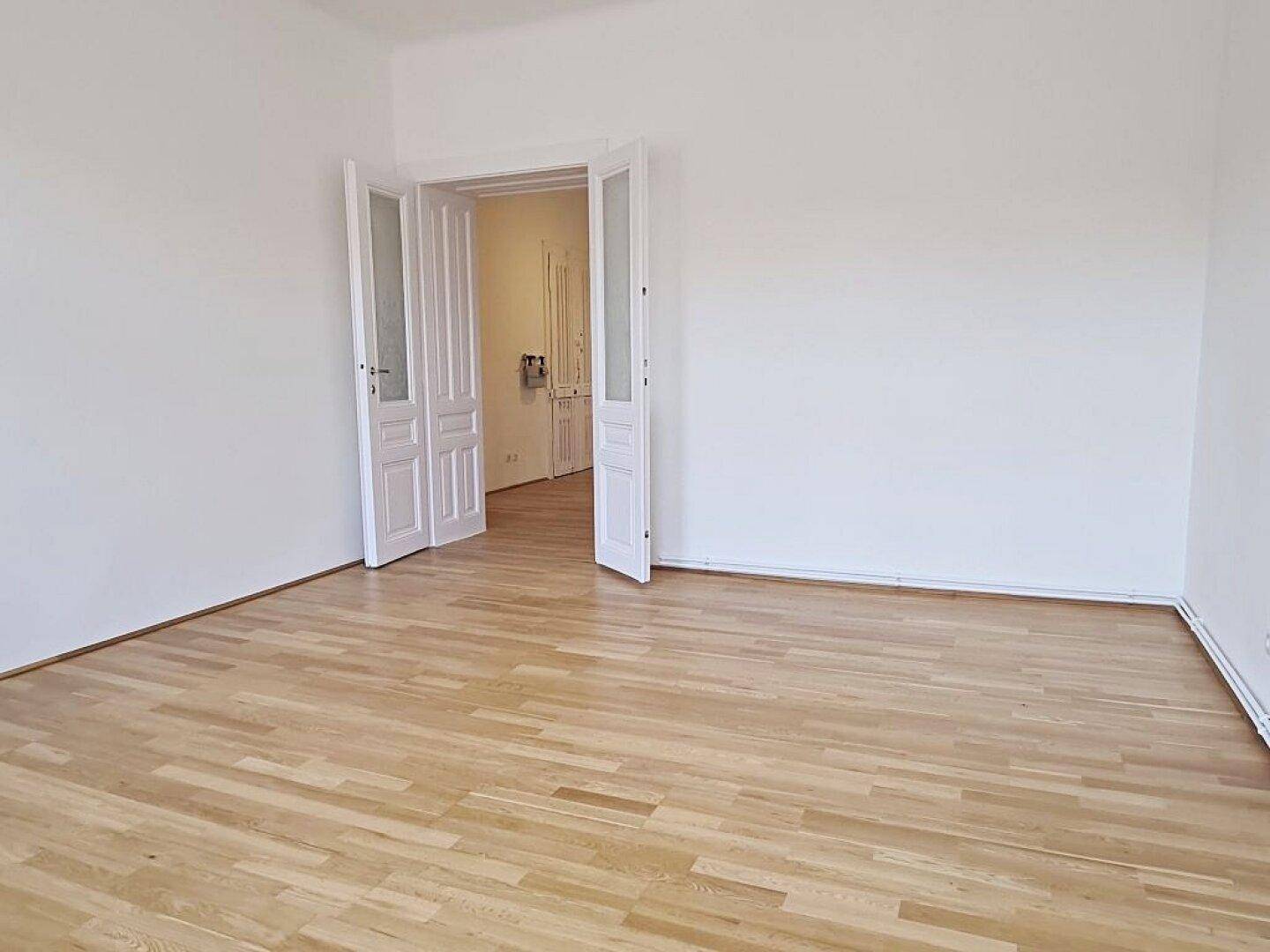 Helles Zimmer mit Parkettboden und klassischen Flügeltüren, die zu einem weiteren Raum führen.