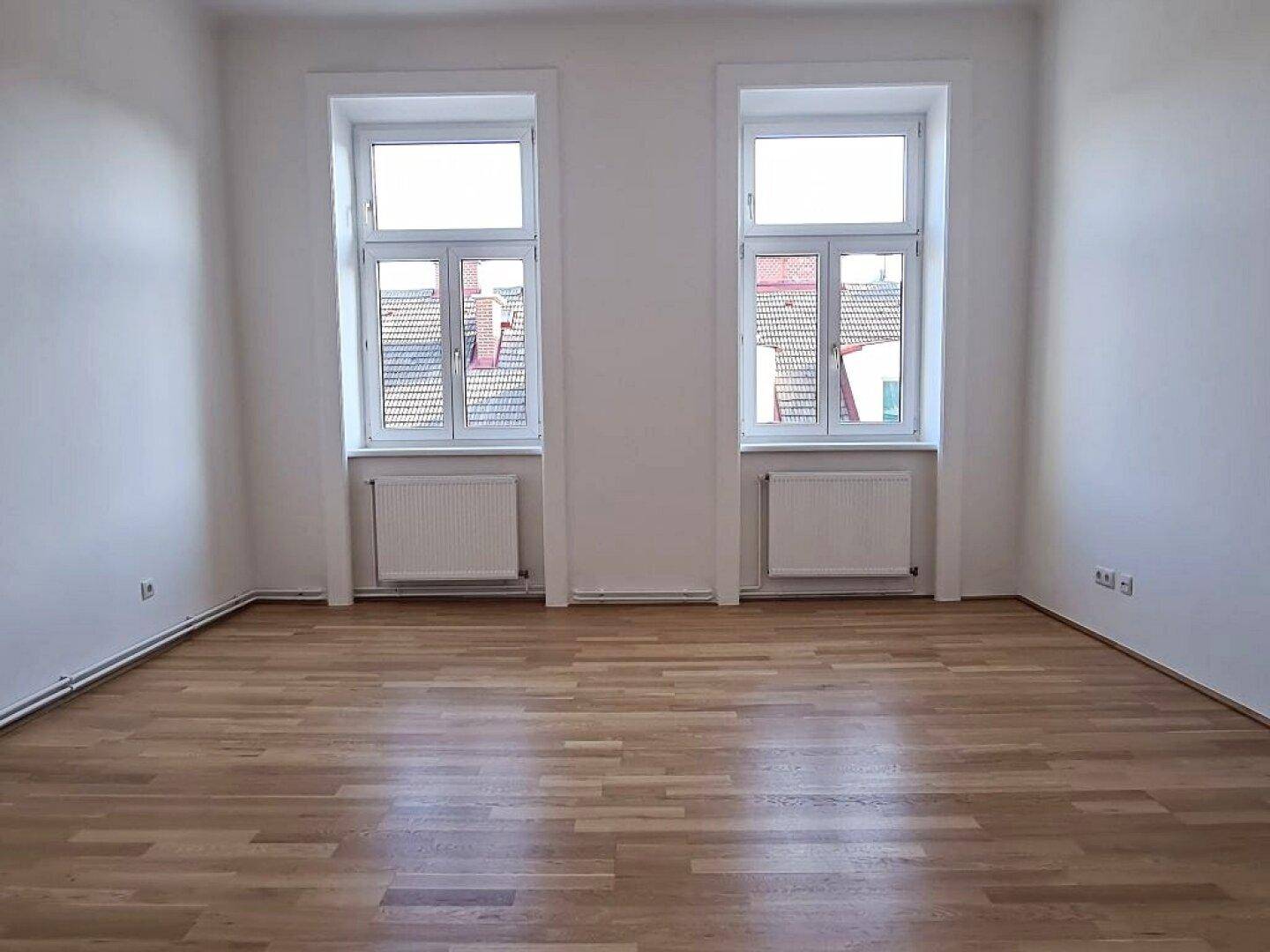 Freundliches Zimmer mit Parkettboden und zwei Fenstern, die viel Tageslicht hereinlassen.