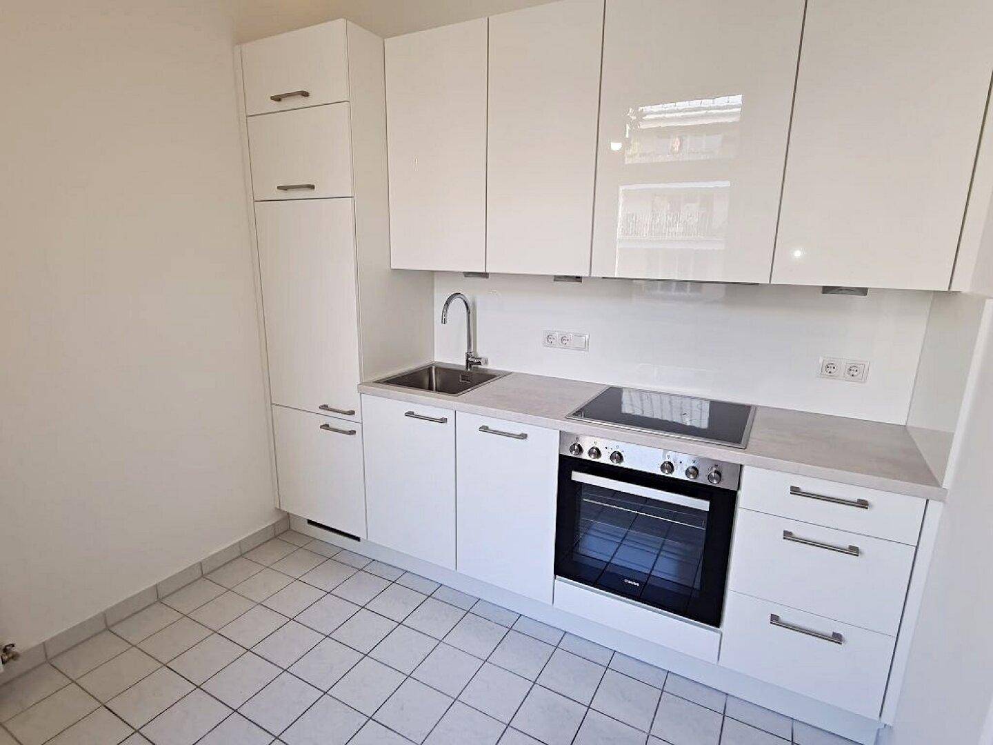 Moderne Einbauküche mit weißen Hochglanzfronten, Induktionskochfeld und Backofen auf Fliesenboden.