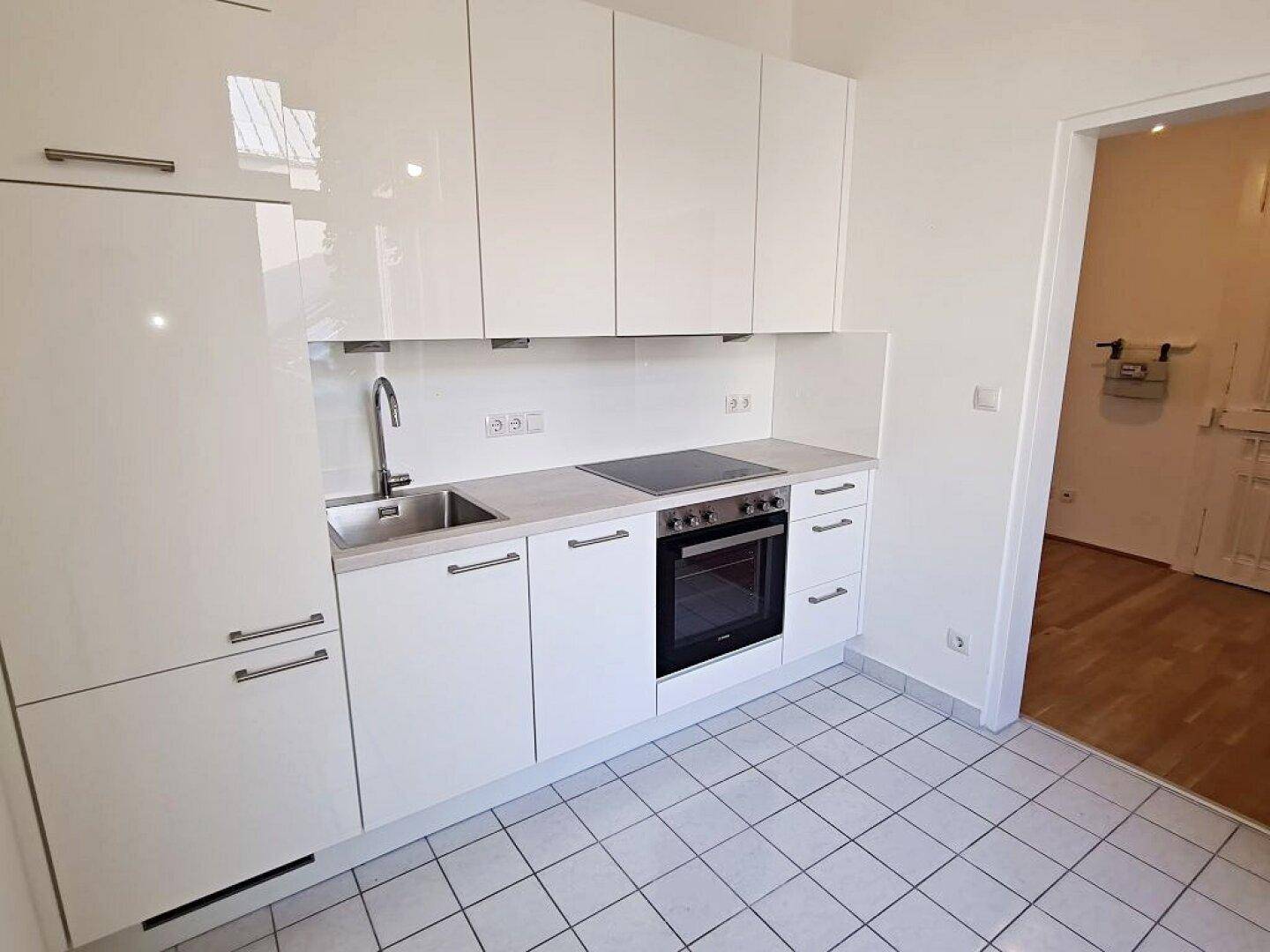 Funktionale Küche mit weißen Hochglanzschränken, Induktionskochfeld und Fliesenboden, offen zum Flur.