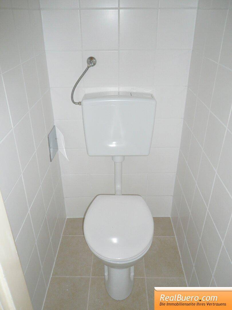 Separates WC mit weißen Wandfliesen und beigefarbenem Boden, ausgestattet mit einer Standardtoilette.
