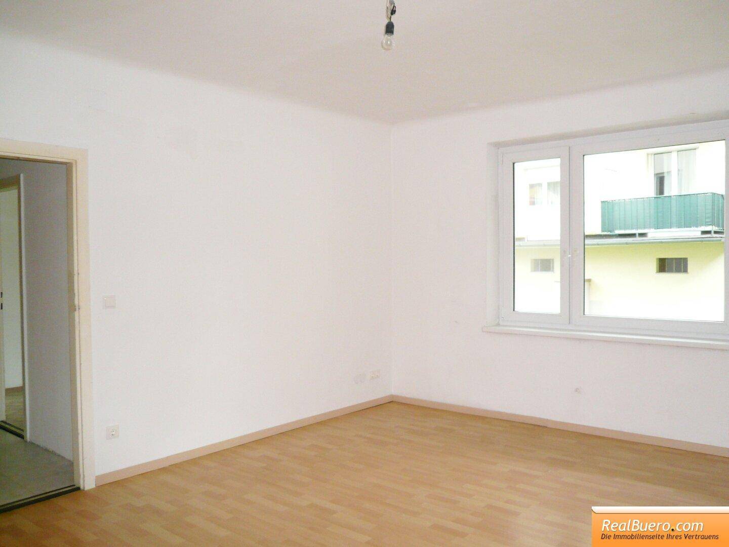 Unmöbliertes Zimmer mit hellem Laminatboden und Fensterblick auf ein benachbartes Gebäude.