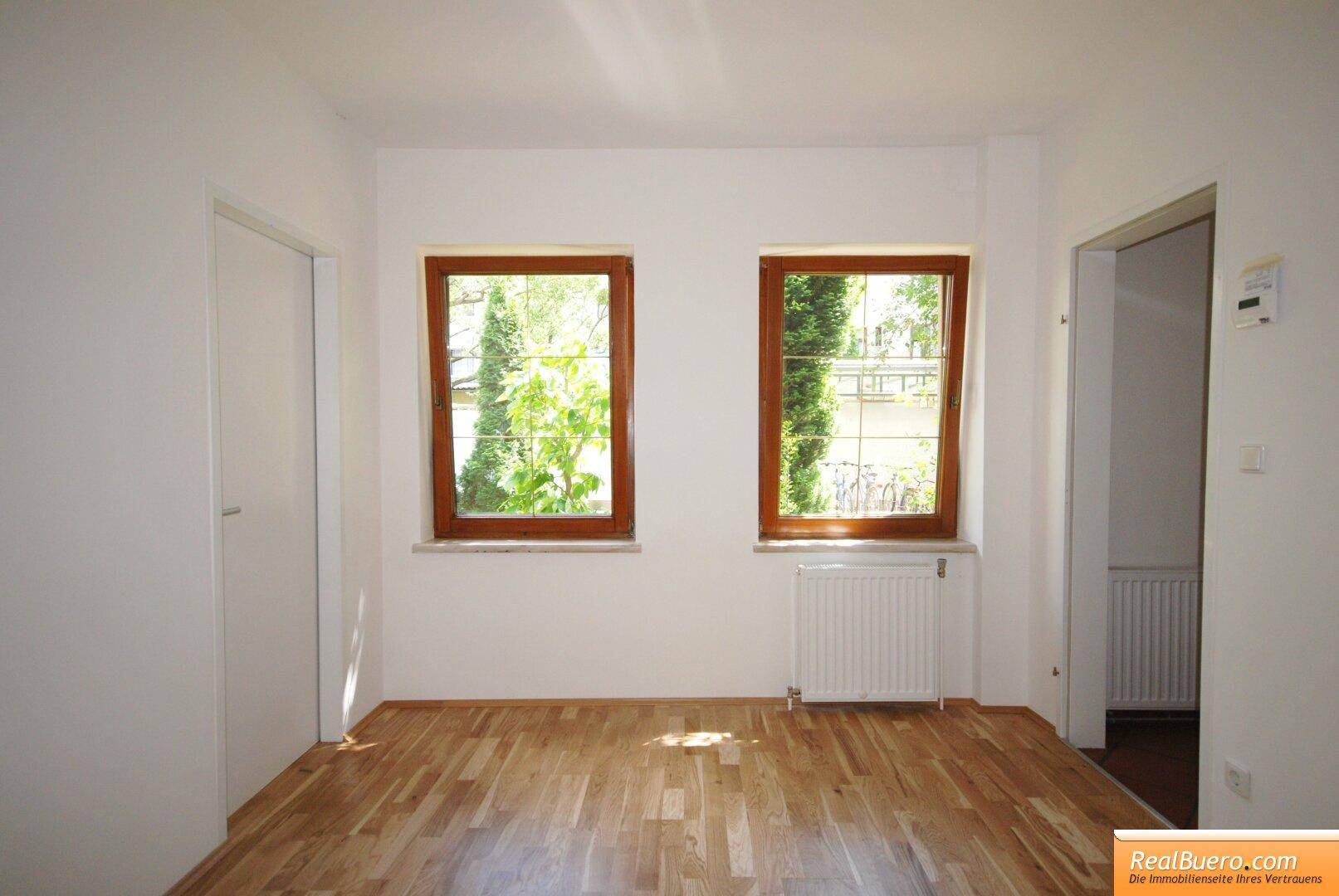 Heller Raum mit Parkettboden und zwei Fenstern, die viel Tageslicht hereinlassen.