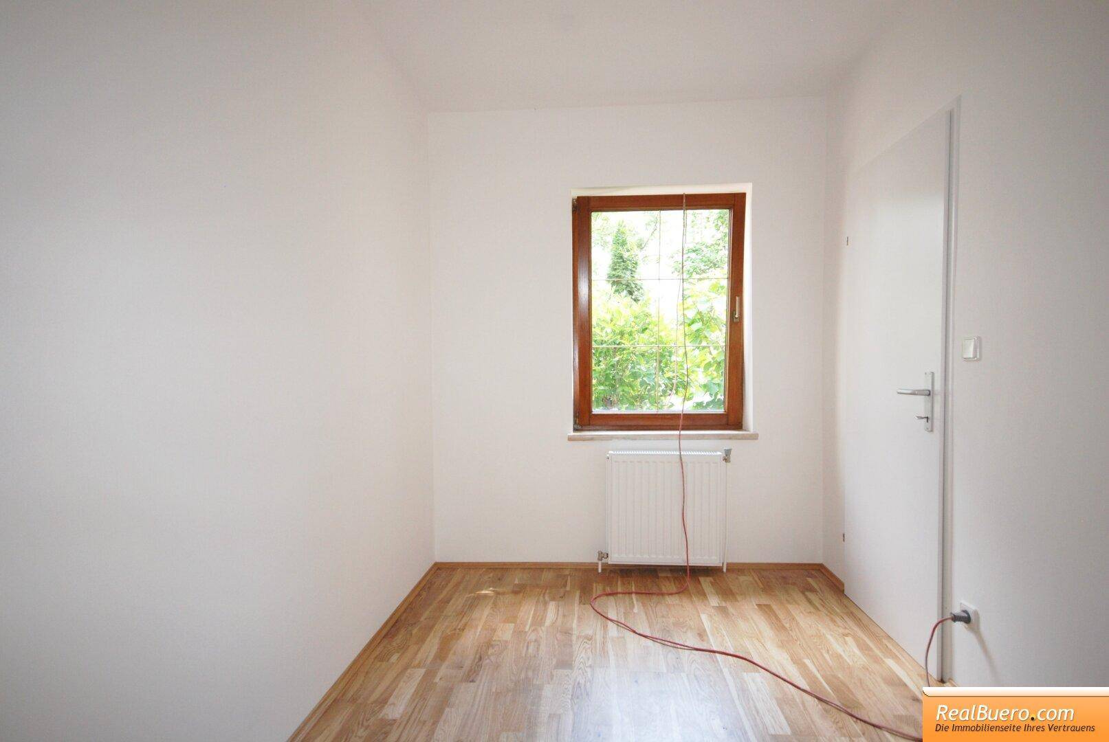 Helles Schlafzimmer mit Parkettboden und einem Fenster mit Blick ins Grüne.