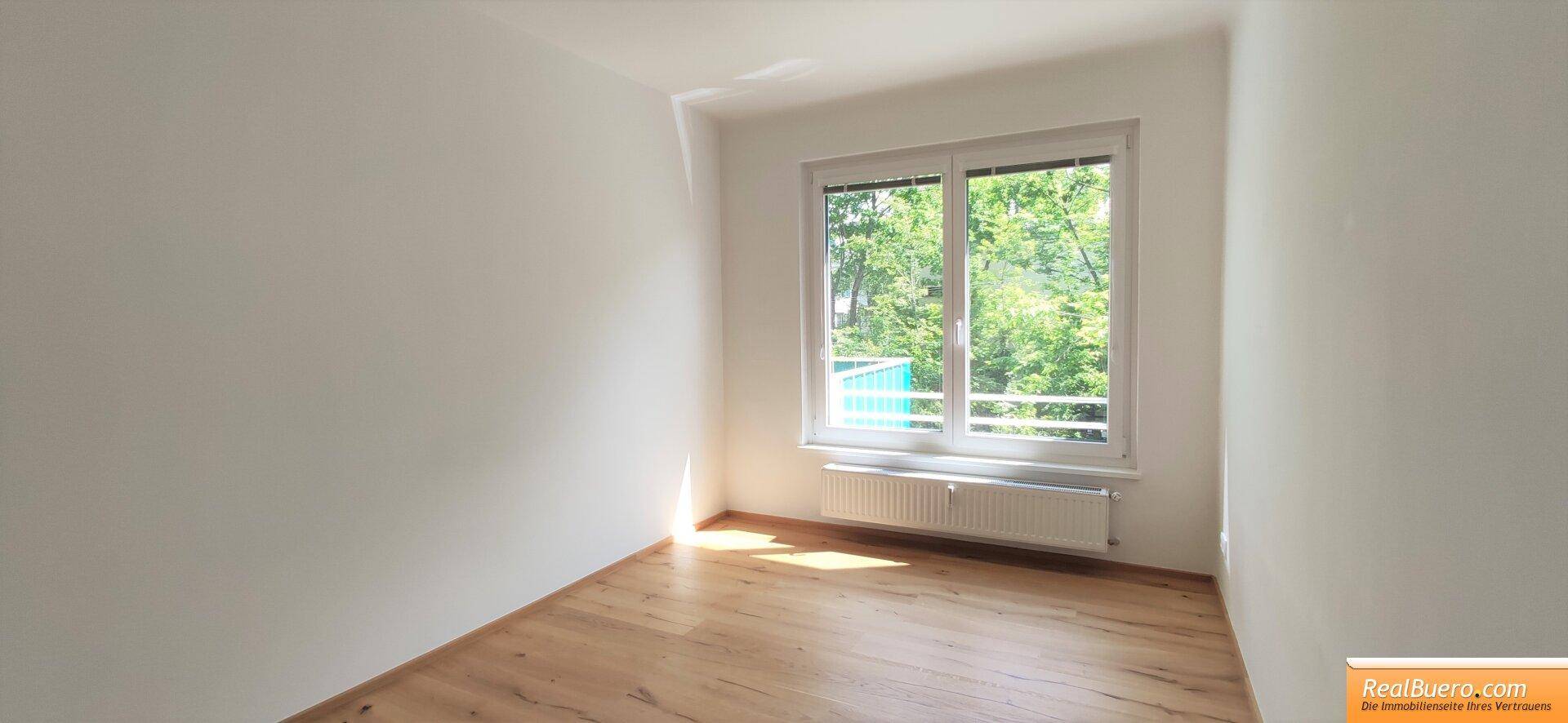 Leeres Zimmer mit Holzboden, großem Fenster und Blick ins Grüne.
