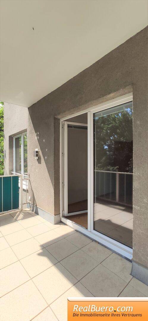 Heller Balkon mit Schiebetürzugang und Fliesenboden, umgeben von Bäumen.