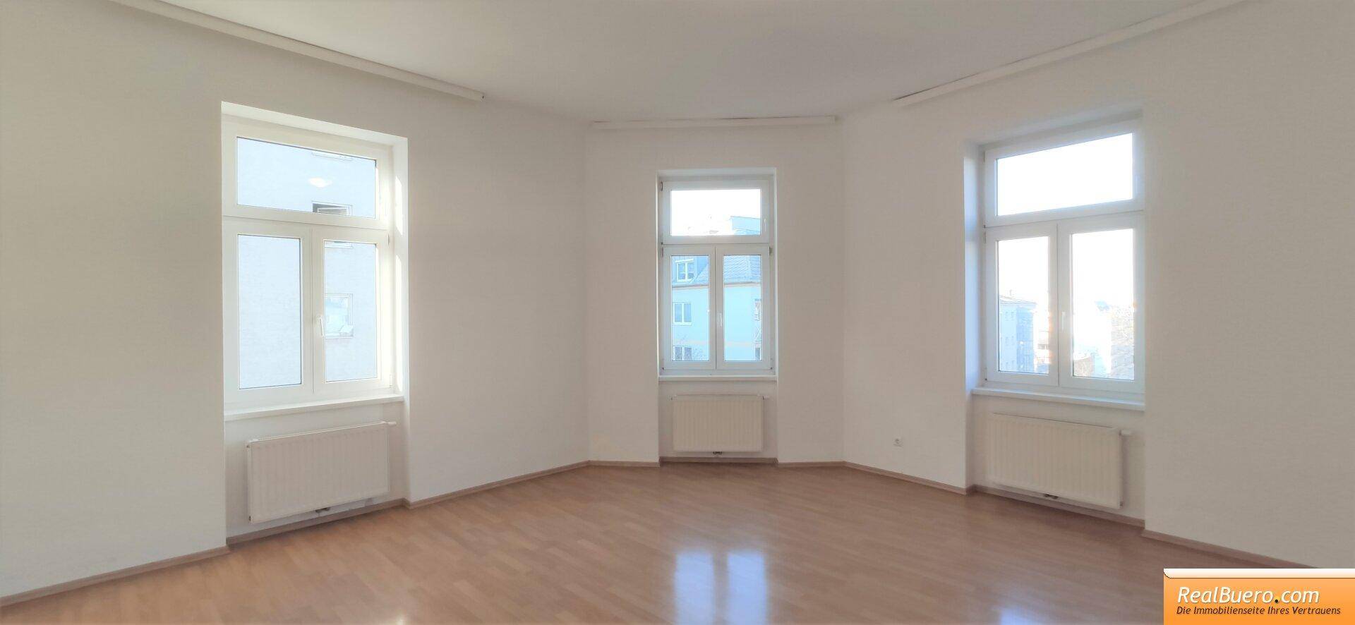 Lichtdurchfluteter Raum mit drei Fenstern, Heizkörpern und einem gepflegten Holzboden.