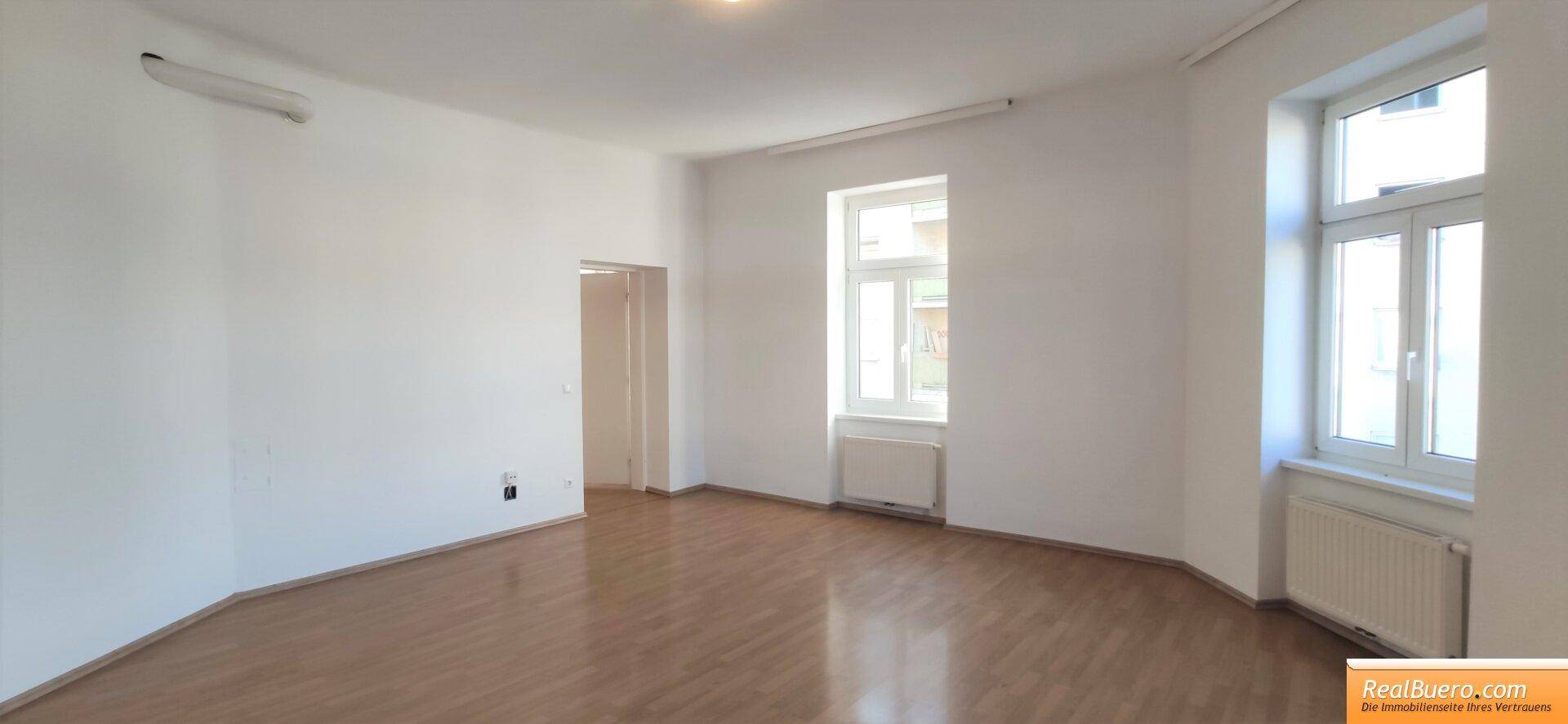 Heller Raum mit Holzboden, zwei Fenstern und einer Klimaanlage an der Wand.