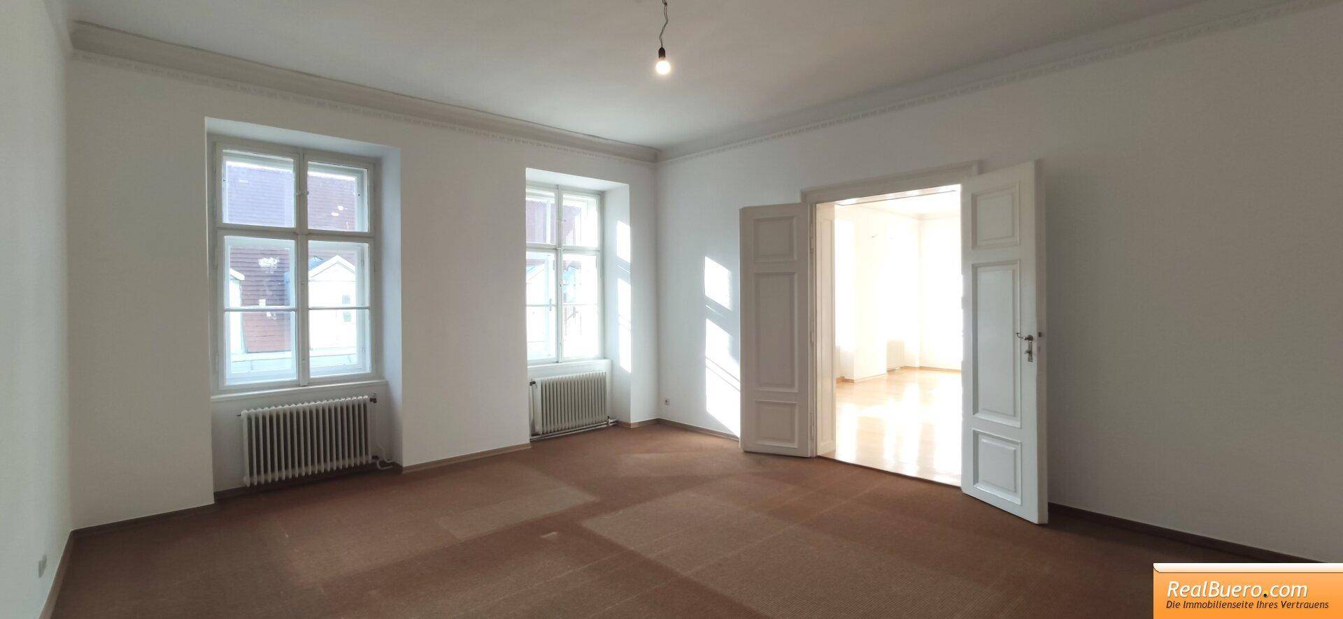 Geräumiger Raum mit braunem Teppichboden, hohen Fenstern und einer Doppeltür zu einem weiteren Zimmer.