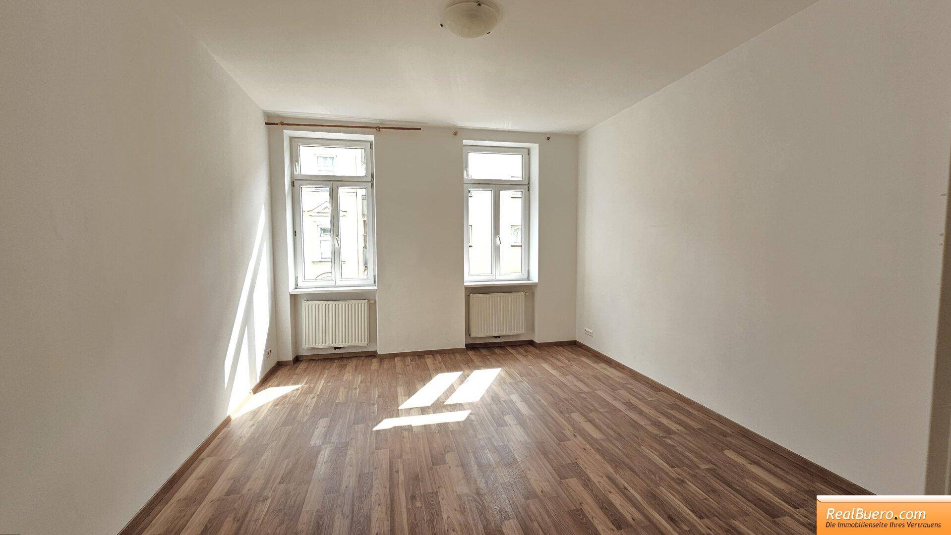 Heller, leerer Raum mit Laminatboden, weißen Wänden und zwei Fenstern, die viel Tageslicht hereinlassen.