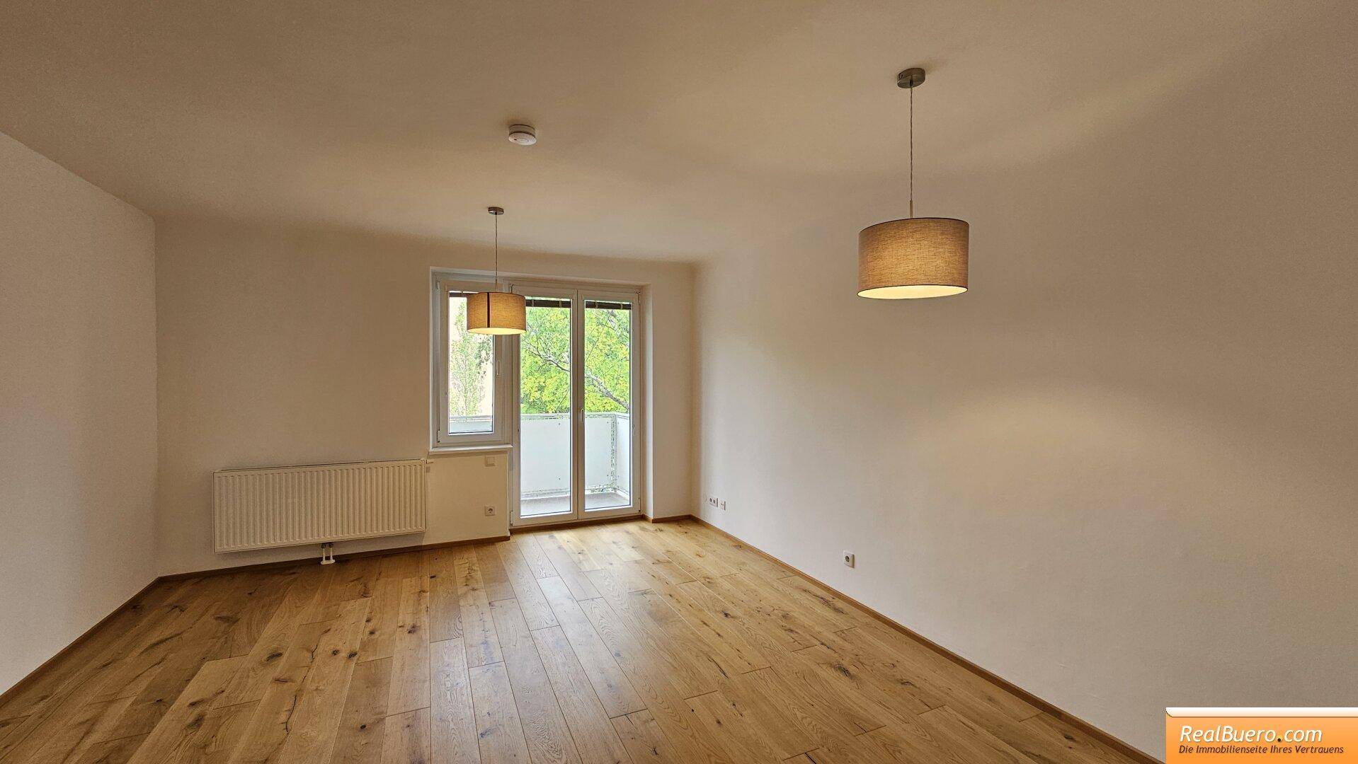 Geräumiges Wohnzimmer mit Holzboden, Heizkörper und Zugang zu einem kleinen Balkon.