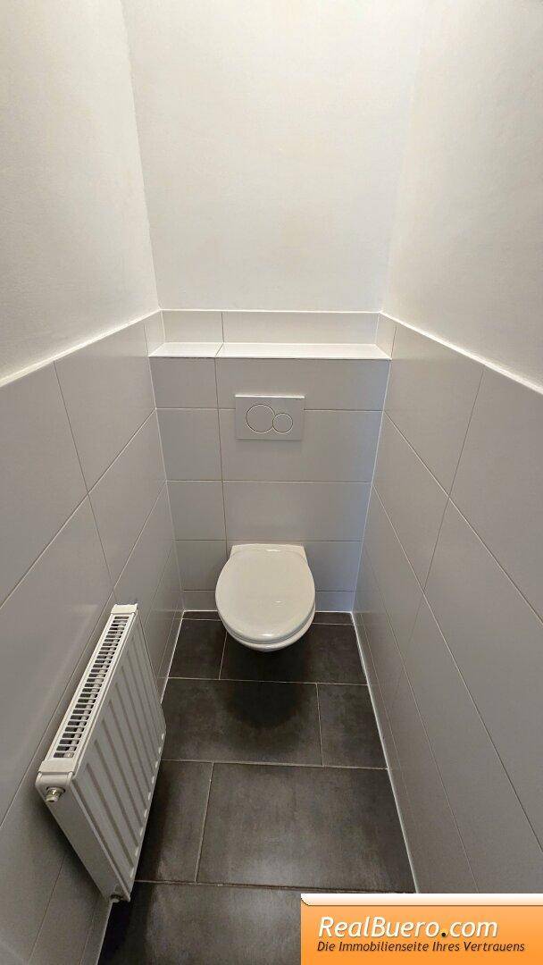 Separates WC mit Wand-WC, Heizkörper und dunklen Bodenfliesen.