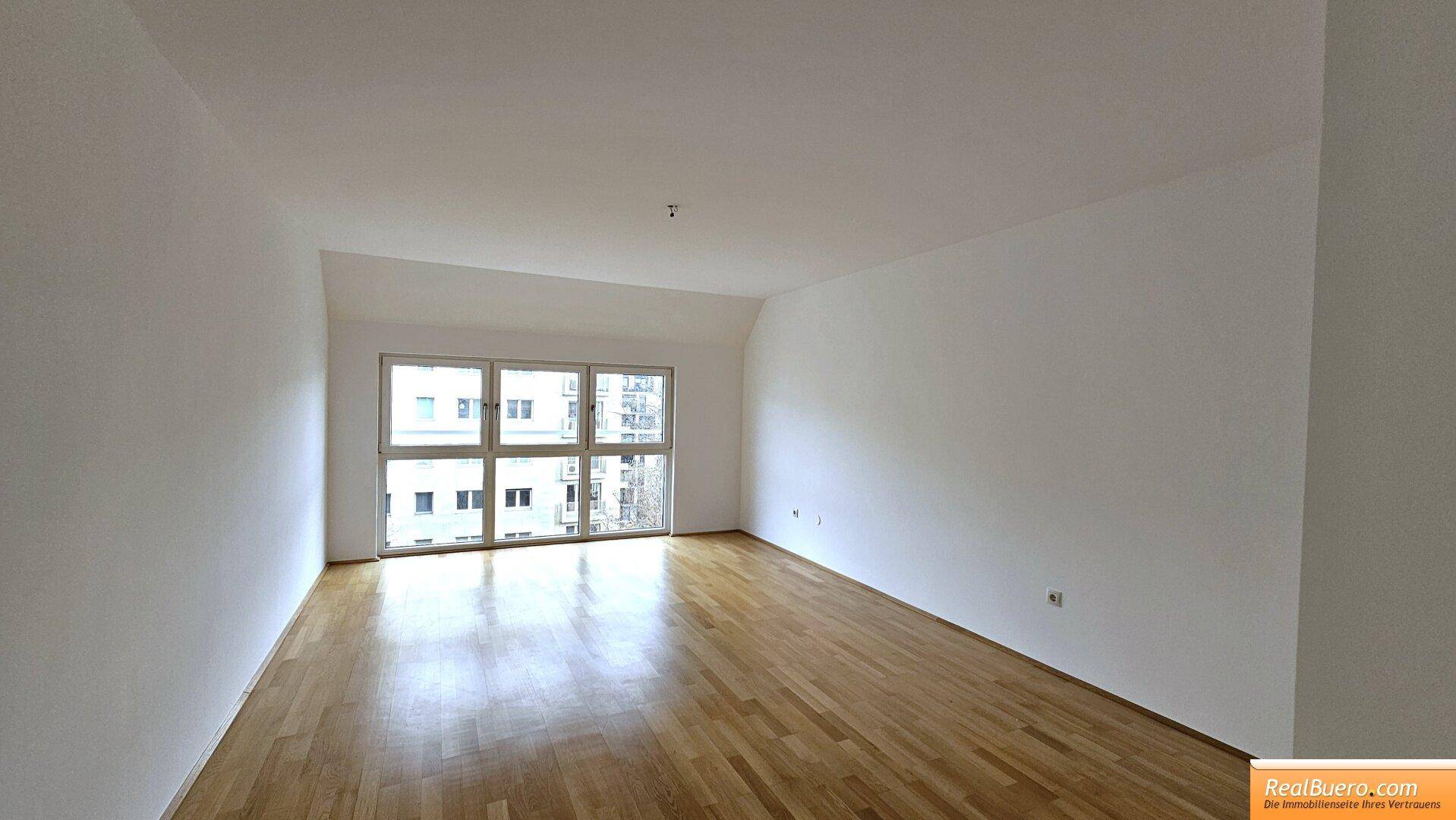 Geräumiges Wohnzimmer mit großen Fenstern, hellem Parkettboden und weißen Wänden.
