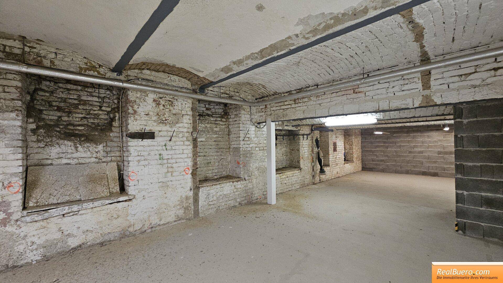 Kellerraum mit unverputzten Ziegelwänden, sichtbaren Rohren und einer gewölbten Decke. Die Beleuchtung ist einfach und zweckmäßig.