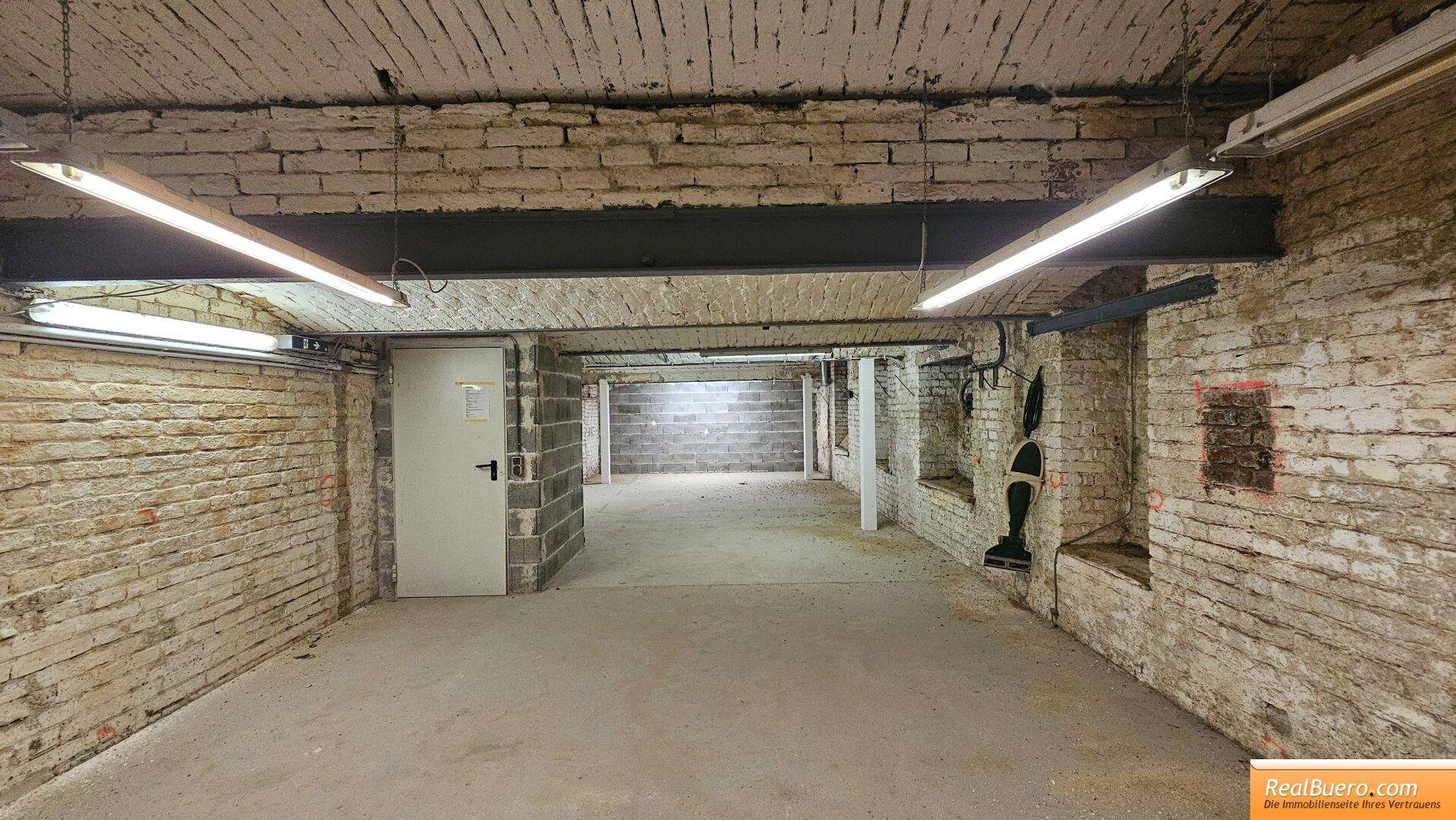 Weiterer Blick in den Kellerbereich mit freiliegenden Ziegeln, Betonboden und funktionaler Deckenbeleuchtung. Die Wände zeigen eine rustikale Textur.