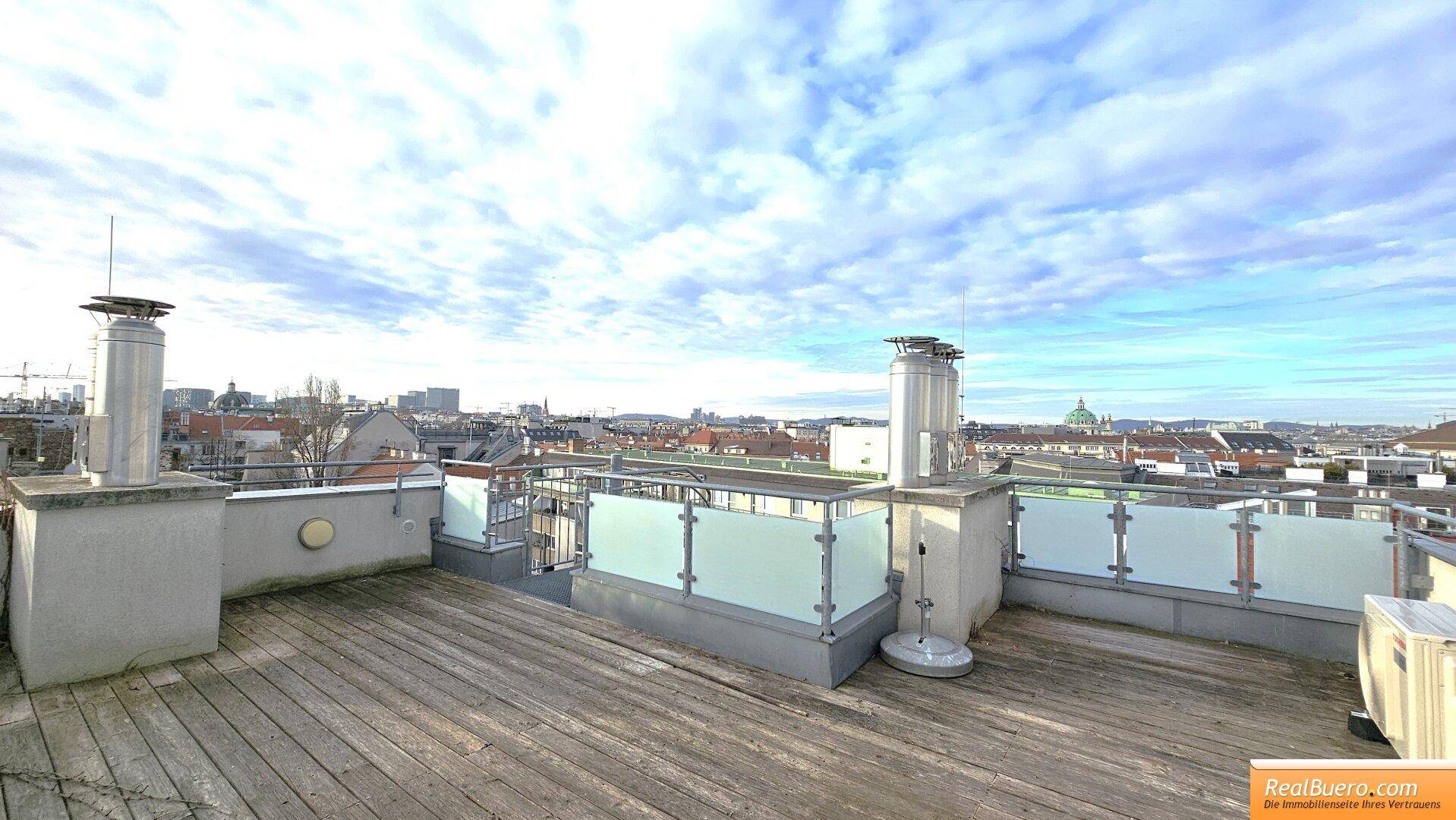 Geräumige Dachterrasse mit Holzboden und weitem Blick über die Stadt unter blauem Himmel.