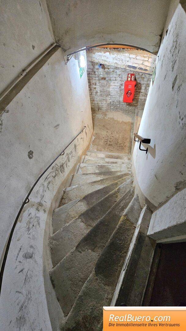 Steile Wendeltreppe führt nach unten, mit einem roten Feuerlöscher an der Wand. Der Handlauf sorgt für Sicherheit.