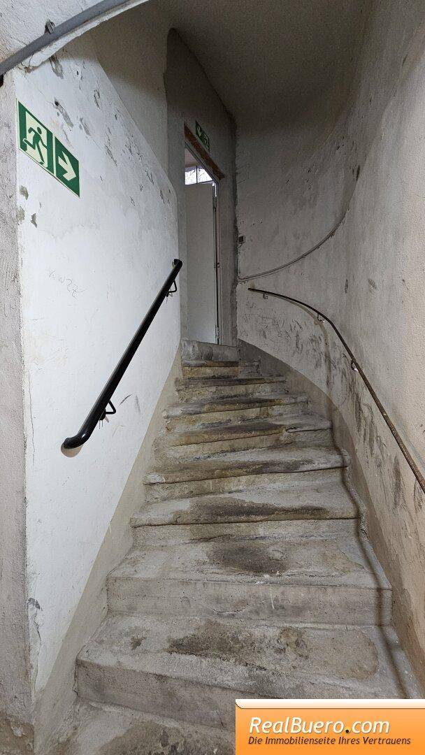Treppenaufgang mit schwarzem Handlauf und Notausgangsschild an der Wand. Die Stufen sind aus robustem Material gefertigt.