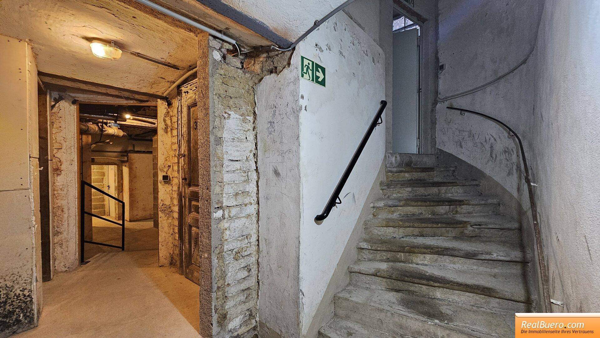 Treppenaufgang mit Handlauf und Notausgangsschild, führt zu einem Flur mit Ziegelwänden. Die Treppe ist aus Stein.