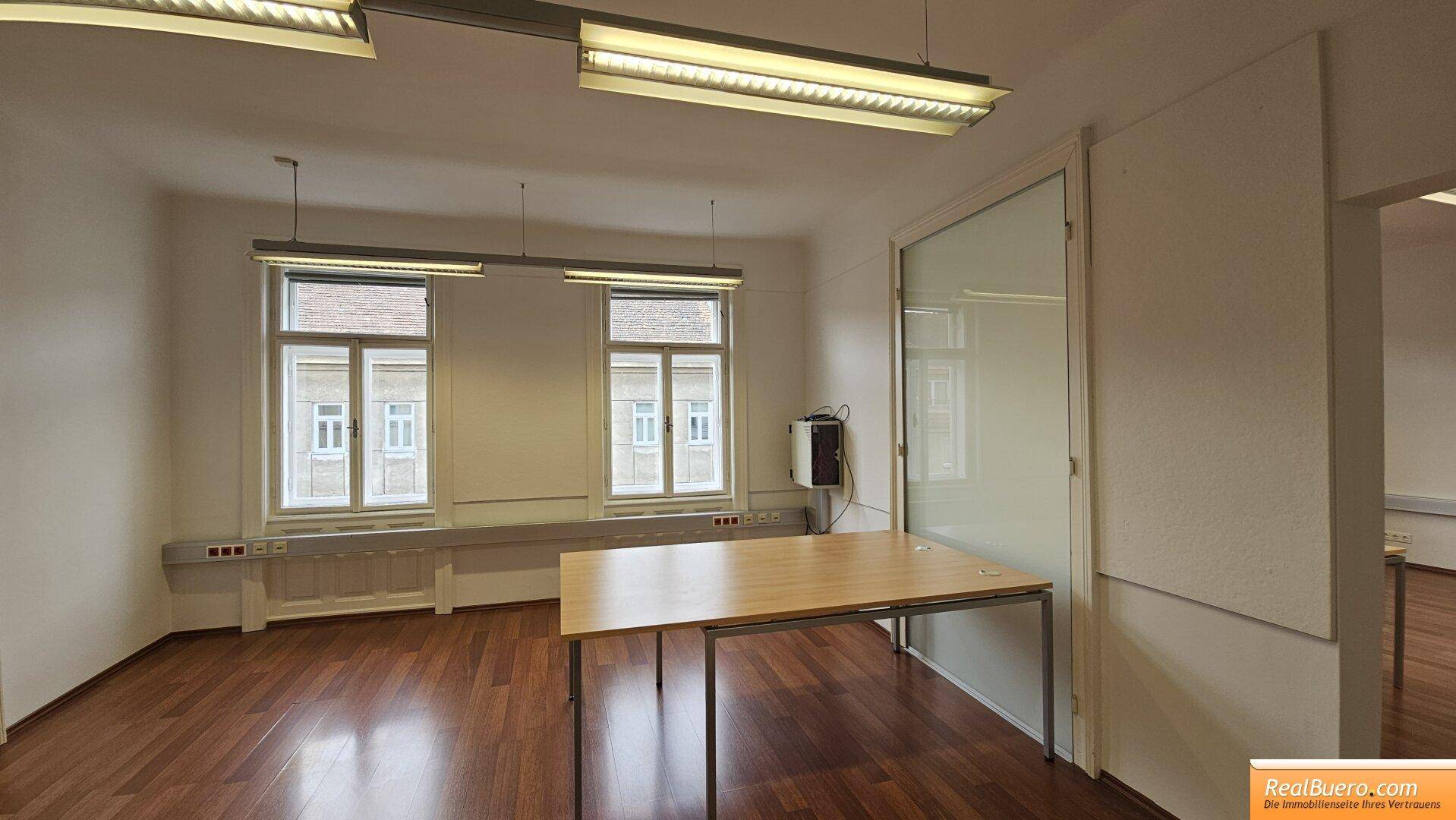 Heller Büroraum mit Holzfußboden, zwei Fenstern und einem modernen Schreibtisch.