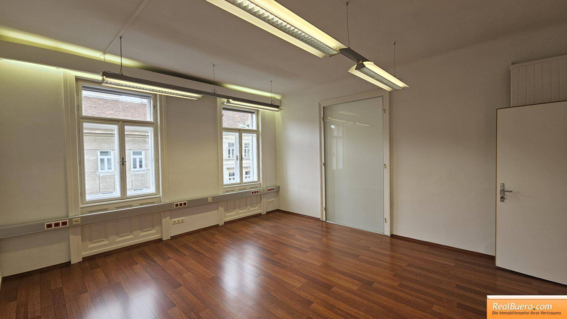 Geräumiger Büroraum mit Holzfußboden, zwei Fenstern und einer Glastür.