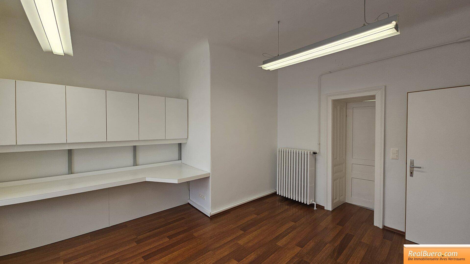 Heller Büroraum mit Holzfußboden, weißen Einbauschränken und Heizkörper.