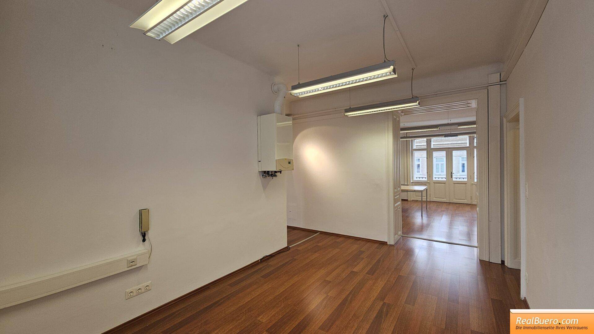 Geräumiger Büroraum mit Holzfußboden, Blick in einen weiteren Raum und Wandheizung.
