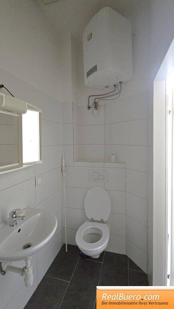 Kleines Badezimmer mit WC, Waschbecken, Wandboiler und weißen Fliesen an den Wänden.