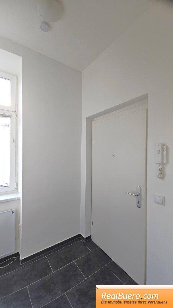 Eingangsbereich mit weißer Tür, Fenster, grauem Fliesenboden und Gegensprechanlage.