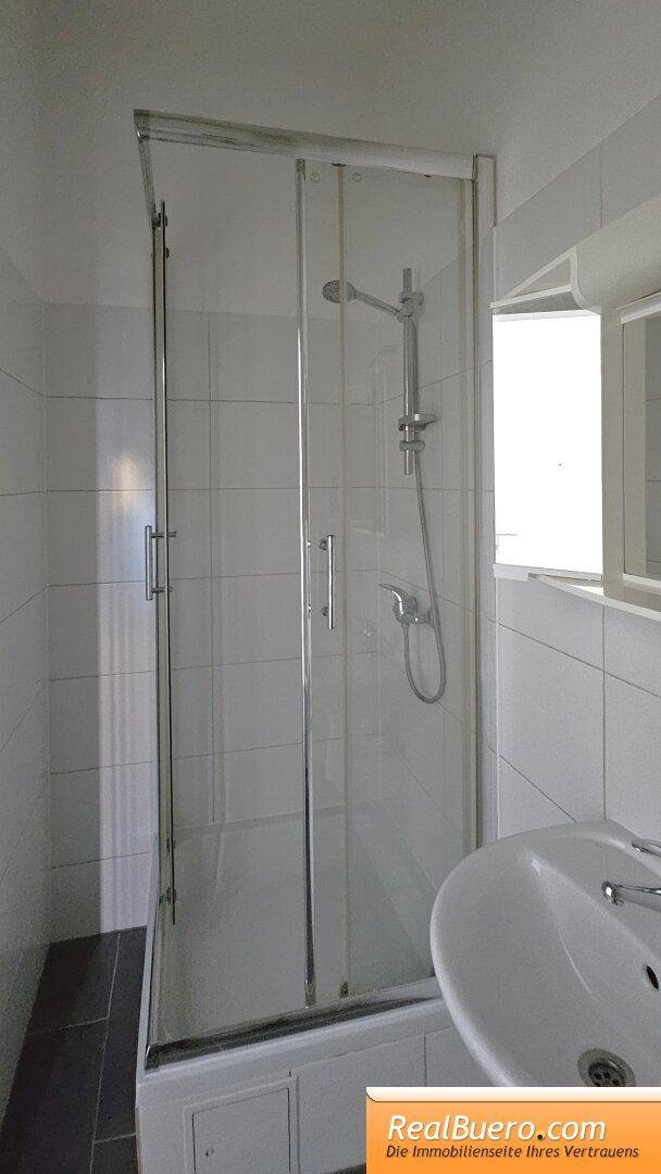 Duschkabine mit Glastüren und Duschkopf neben einem Waschbecken in einem gefliesten Bad.