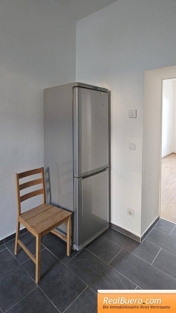 Freistehender Kühlschrank aus Edelstahl neben einem Holzstuhl auf grauem Fliesenboden.