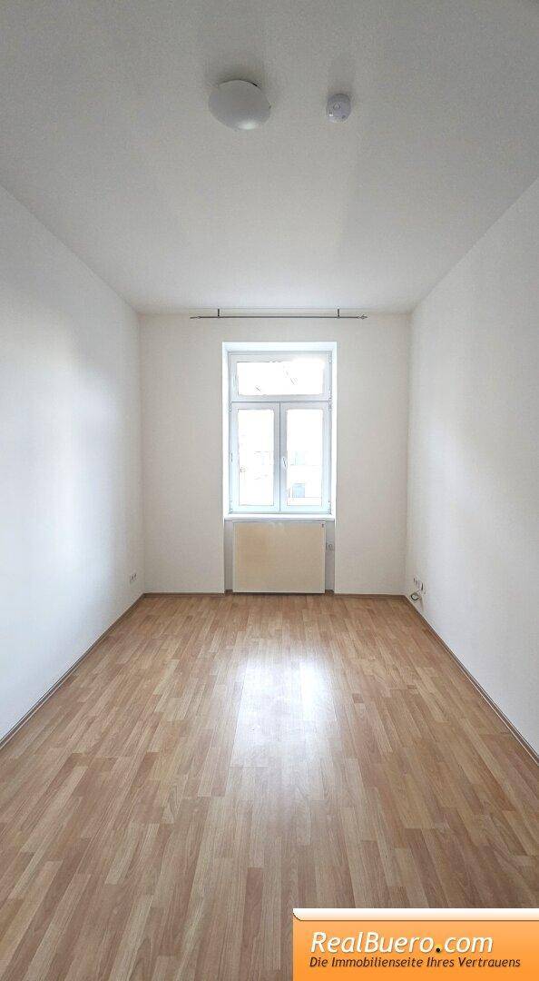 Leeres Zimmer mit hellem Laminatboden, weißen Wänden und einem Fenster mit Heizkörper.