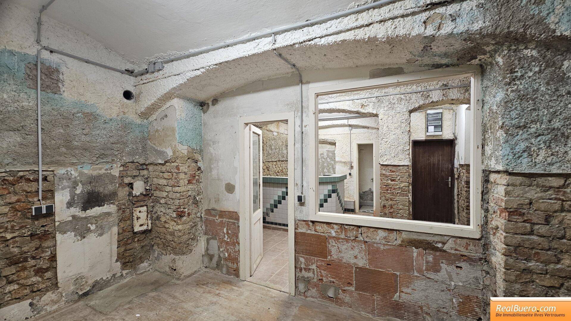 Kellerraum mit unverputzten Wänden, einem Fenster und einem Durchgang zu einem Treppenbereich, der Renovierung benötigt.