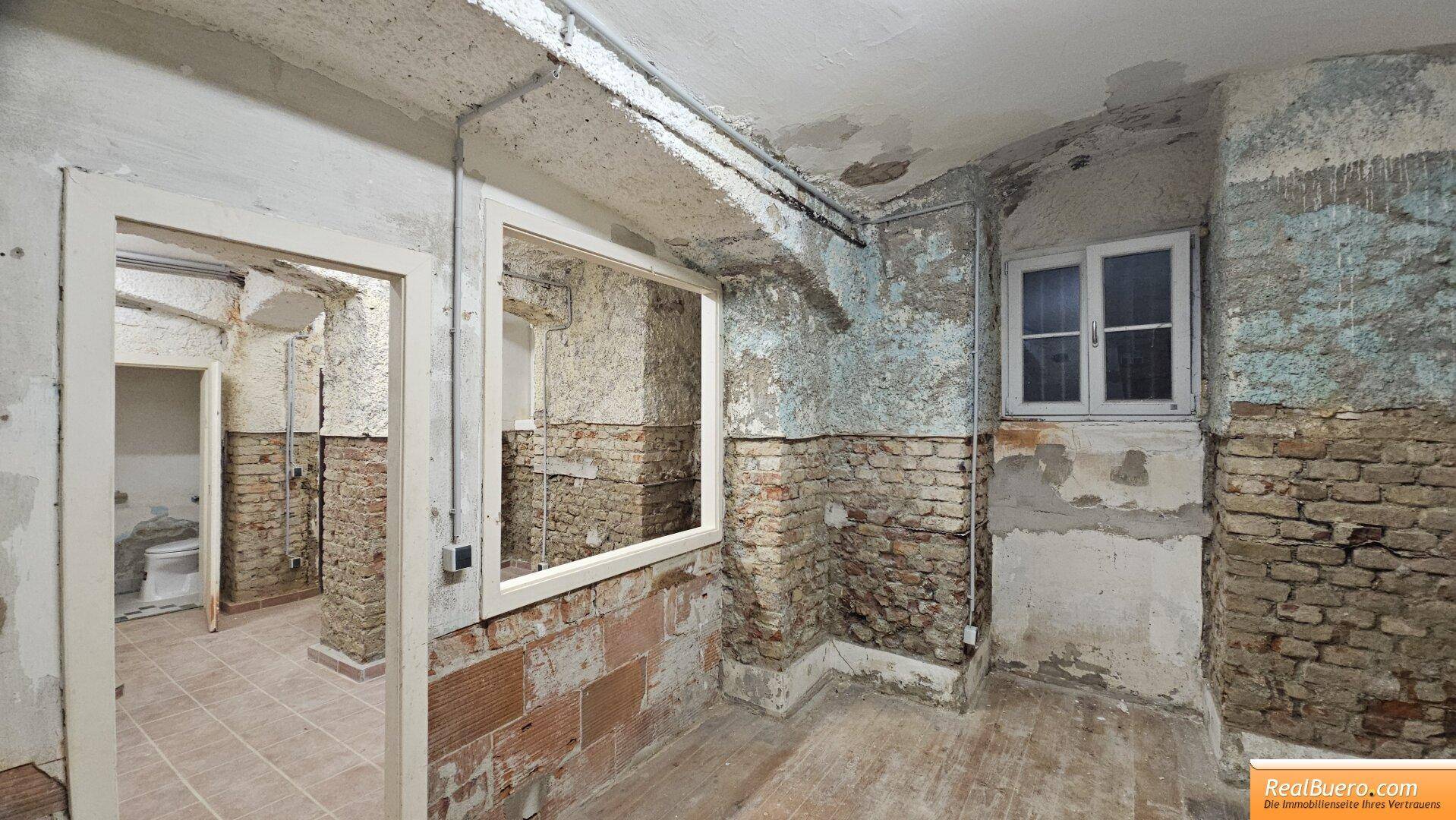 Raum mit unverputzten Ziegelwänden, Fenster und Zugang zu einem WC-Bereich, mit gemischtem Bodenbelag.