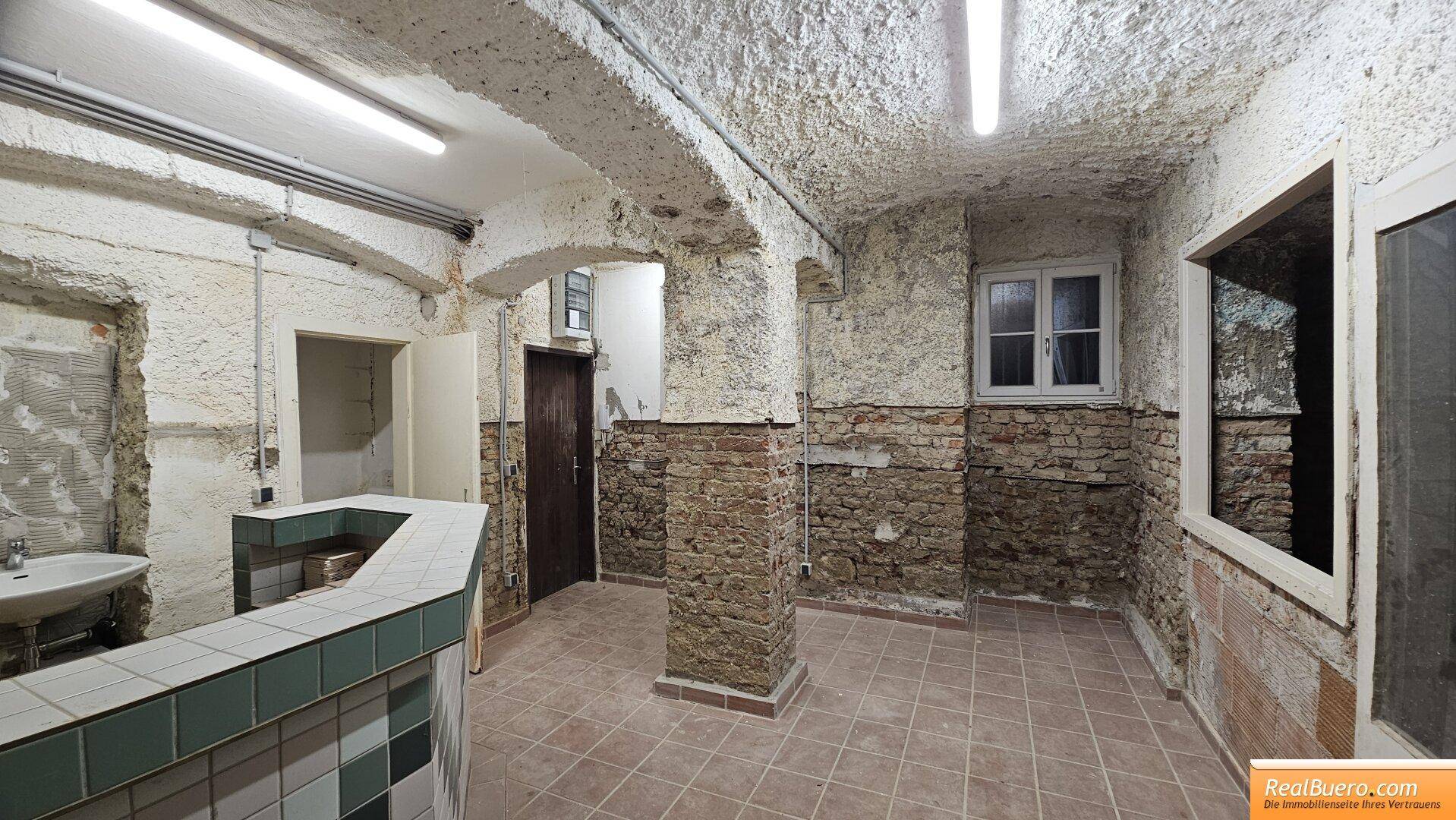 Kellerraum mit gefliester Theke, Waschbecken und Fenster, umgeben von unverputzten Ziegelwänden und rauer Decke.