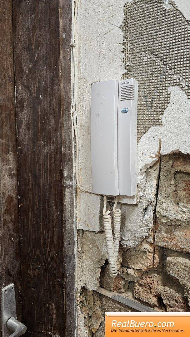 Gegensprechanlage neben einer Holztür an einer unverputzten Wand, die den Eingangsbereich ergänzt.