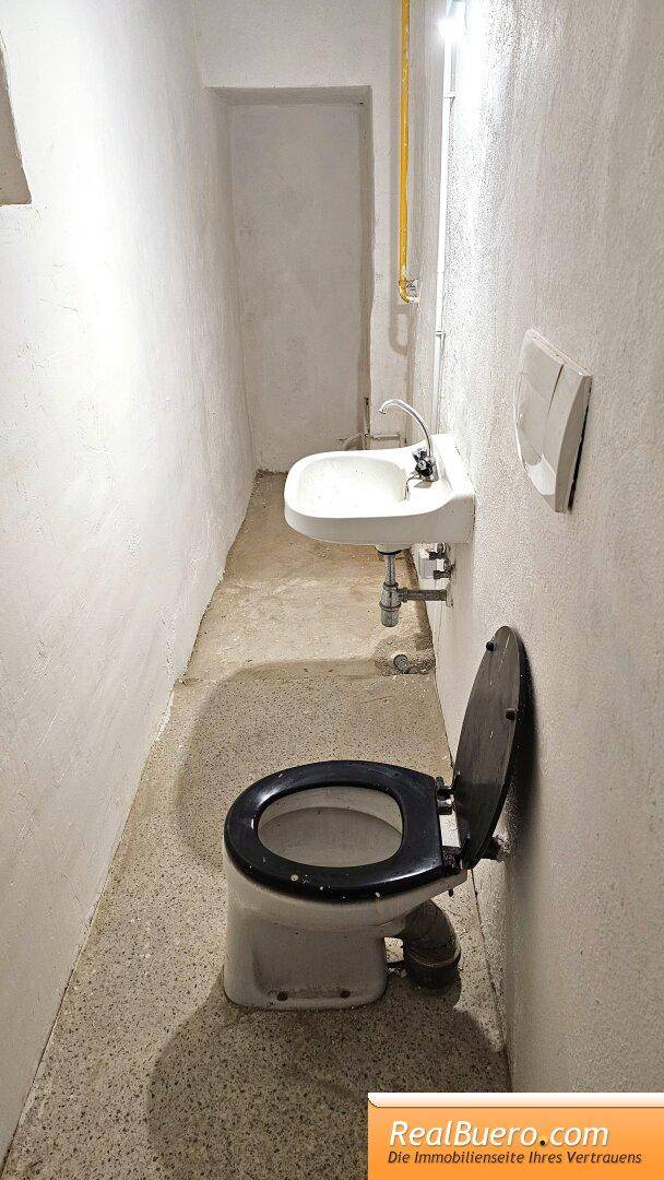 Einfache Toilette mit Waschbecken und sichtbaren Leitungen in einem schmalen Raum.