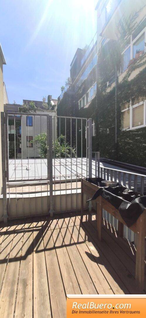 Heller Gartenbereich mit Holzboden und Metallzaun, umgeben von begrünten Fassaden.