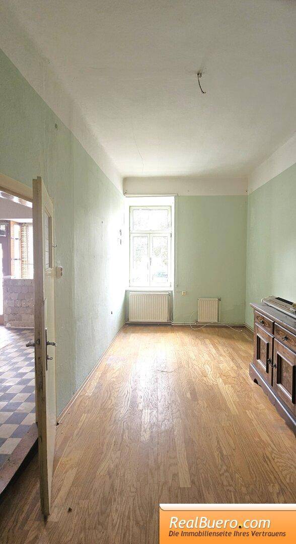 Helles Zimmer mit Holzboden, hellgrünen Wänden und einem Fenster mit Blick nach draußen.