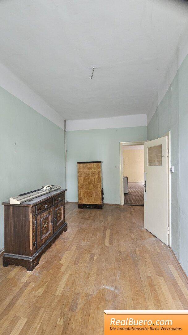 Geräumiges Zimmer mit hellgrünen Wänden, Holzboden und einem traditionellen Kachelofen.