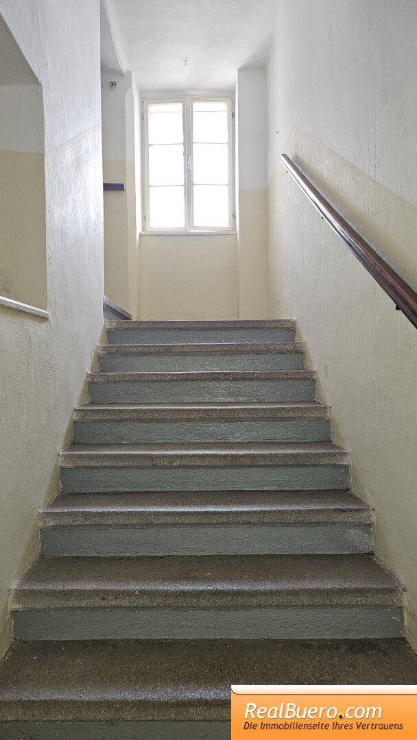 Treppenhaus mit Steinstufen und Holzgeländer, führt zu einem hellen Fenster im Obergeschoss.