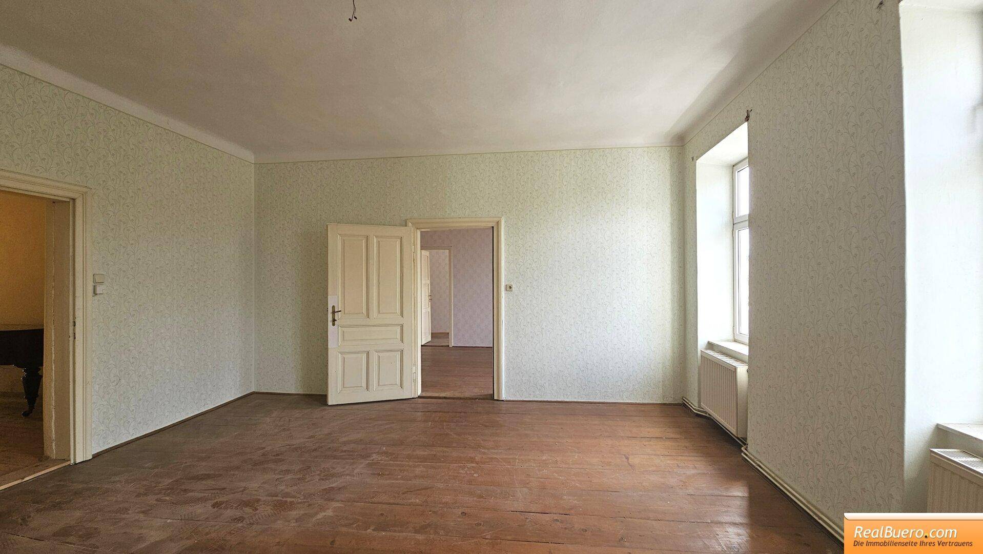 Großes Zimmer mit Holzdielenboden und gemusterter Tapete, zwei Fenstern und einer Tür.