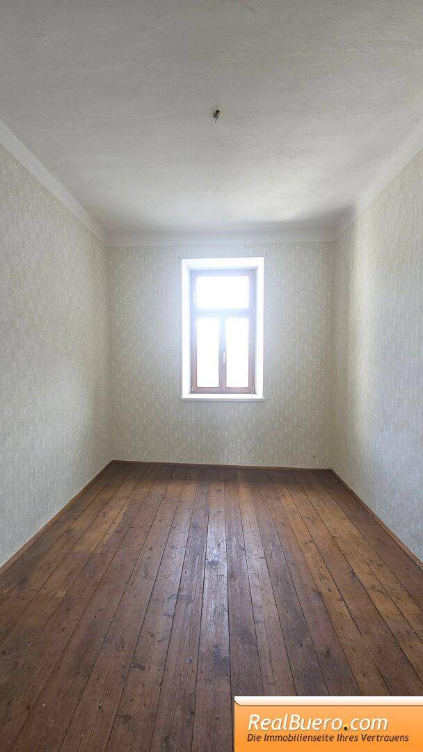 Helles Zimmer mit Holzdielenboden und gemusterter Tapete, einem Fenster und viel Tageslicht.