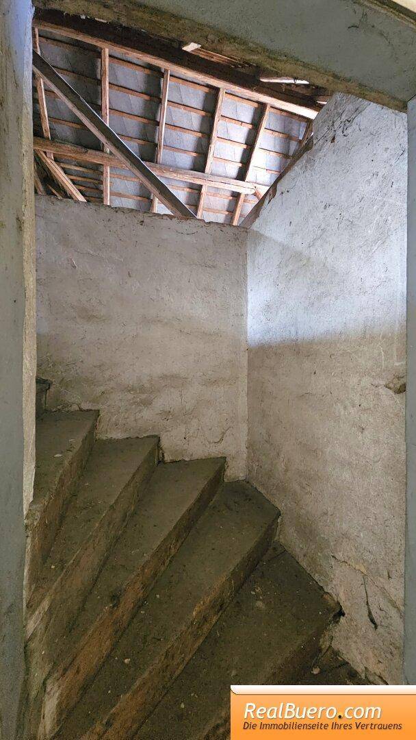 Treppe führt zu einem unfertigen Dachboden mit sichtbaren Holzbalken und rauen Wänden.