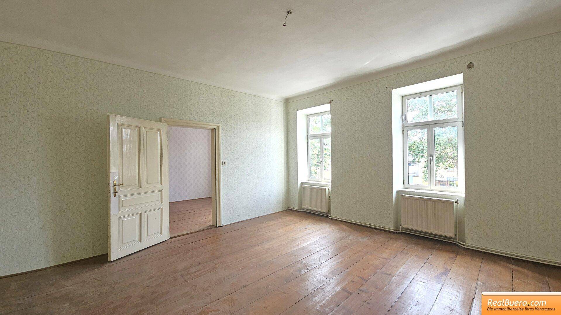 Geräumiges Wohnzimmer mit Holzdielenboden und zwei großen Fenstern, bereit zur Einrichtung.