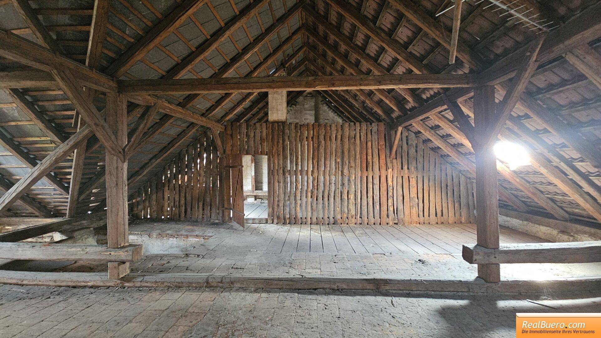 Unfertiger Dachboden mit Holzdielenboden, sichtbaren Balken und einer einfachen Holztrennwand.
