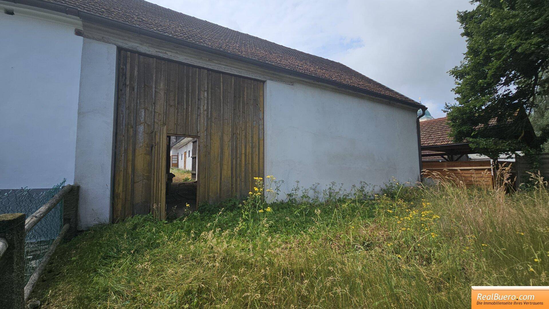 Seitenansicht eines alten Gebäudes mit großer Holztür und hohem Gras im Vordergrund.