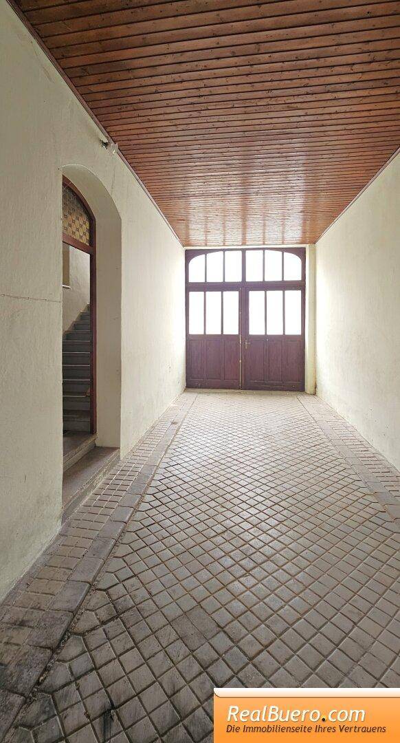 Heller Flur mit Holztäfelung an der Decke und gefliestem Boden, führt zu einer großen Holztür.