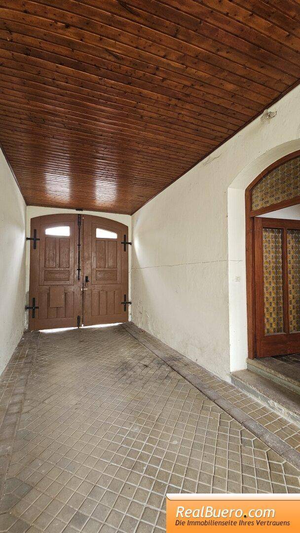 Langer Durchgang mit Holztäfelung an der Decke und gefliestem Boden, führt zu einer Holztür.