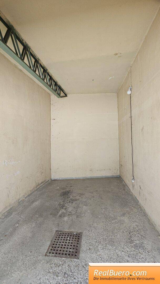 Leerer Lagerraum mit Betonboden, einem Abfluss und einfacher Beleuchtung an der Wand.